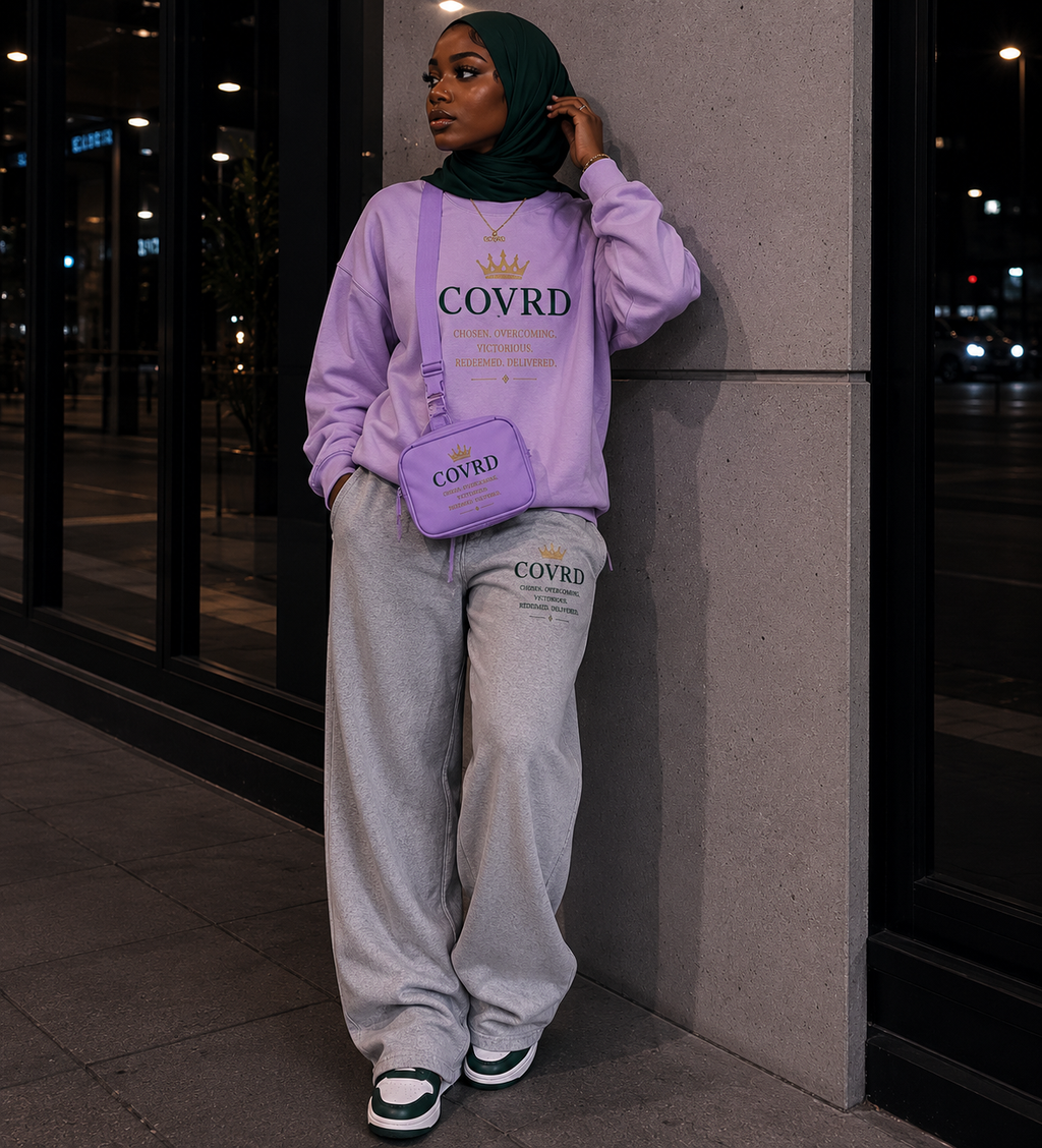 Person wearing a light pink sweatshirt and gray pants with sling bag with 'COVRD' branding, standing against a building.