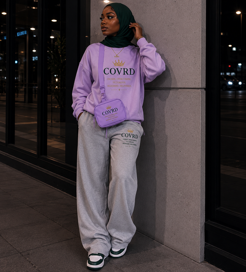Person wearing a light pink sweatshirt and gray pants with sling bag with 'COVRD' branding, standing against a building.